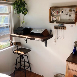 May include: A home office setup featuring a wooden desk with a vintage typewriter and laptop. A metal and wood adjustable stool sits in front of the desk. Jewelry is displayed on the wall, and a hanging plant adds a natural touch.