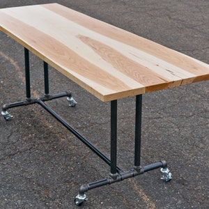 May include: A rectangular wooden table with a light brown surface and black metal pipe legs. The table is supported by four legs with caster wheels. The tabletop is approximately 60 inches long.