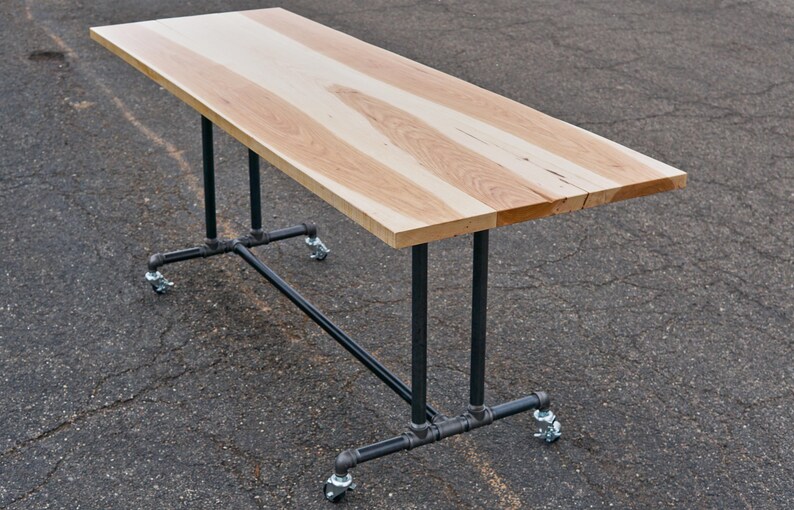 May include: A rectangular wooden table with a light brown surface and black metal pipe legs. The table is supported by four legs with caster wheels. The tabletop is approximately 60 inches long.