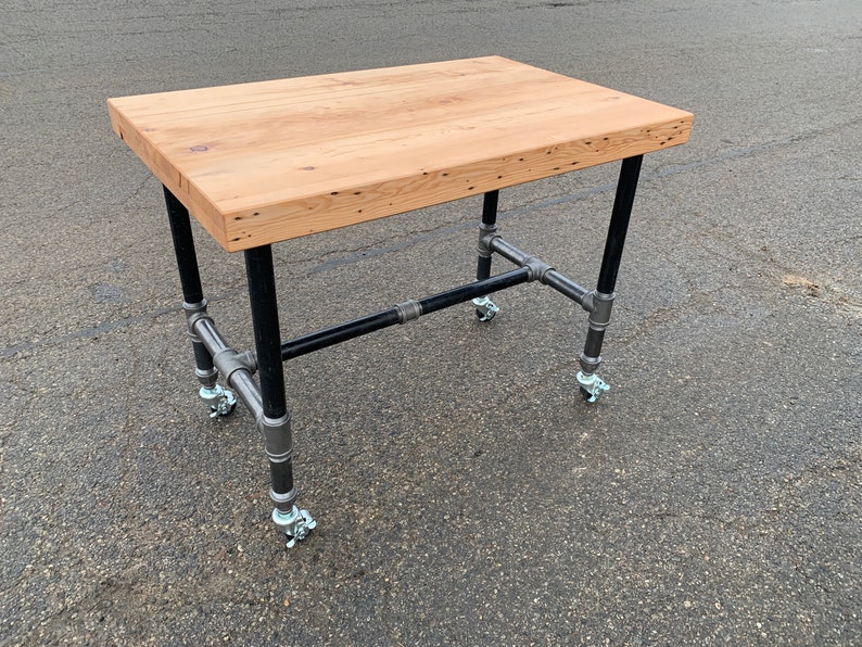 May include: A rectangular wooden table with a light brown finish. The table has black metal pipe legs and a metal pipe frame. The table is 76 cm long, 61 cm wide, and 91 cm high. The table has four caster wheels.