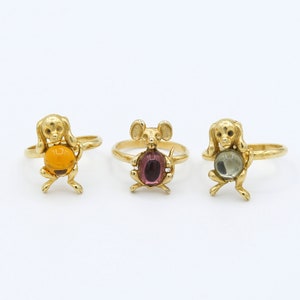 May include: Three gold rings with animal figures holding colorful gemstones. The rings feature a dog holding an amber stone, a mouse holding a purple stone, and a dog holding a green stone.