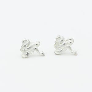 May include: A pair of silver-colored frog-shaped stud earrings. The earrings are detailed with textured surfaces, showcasing the form of a frog. The frog's legs are extended, and the design is set against a white background.