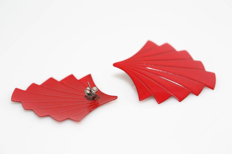 May include: A pair of red fan-shaped earrings with silver posts. The earrings are made of metal and have a glossy finish.