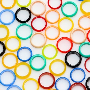 May include: A collection of colorful rings made of smooth, polished stone. The rings are arranged in a random pattern on a white background. The rings are various colors, including red, orange, yellow, green, blue, purple, and black.
