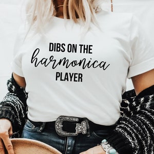 May include: White t-shirt with black text that reads "DIBS ON THE harmonica PLAYER". The person is wearing a black and white striped cardigan, blue jeans, and a brown hat.