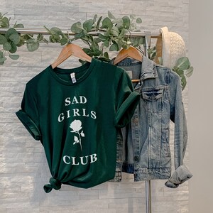 May include: A dark green t-shirt with the text "SAD GIRLS CLUB" and a white rose graphic. The shirt is hanging on a rack with a denim jacket.