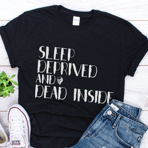 K&ouml;nnte beinhalten: Schwarzes T-Shirt mit wei&szlig;em Text, der "Sleep Deprived and Dead Inside" lautet. Der Text ist in einer handschriftlichen Schriftart gehalten. Das Shirt liegt auf einer wei&szlig;en Holzoberfl&auml;che mit einem Paar wei&szlig;en Sneakers und einer kleinen Topfpflanze.