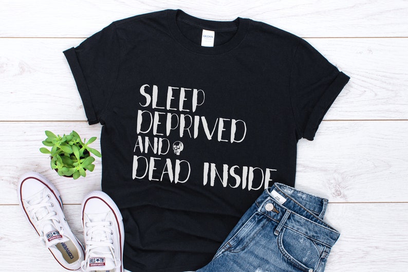 K&ouml;nnte beinhalten: Schwarzes T-Shirt mit wei&szlig;em Text, der "Sleep Deprived and Dead Inside" lautet. Der Text ist in einer handschriftlichen Schriftart gehalten. Das Shirt liegt auf einer wei&szlig;en Holzoberfl&auml;che mit einem Paar wei&szlig;en Sneakers und einer kleinen Topfpflanze.