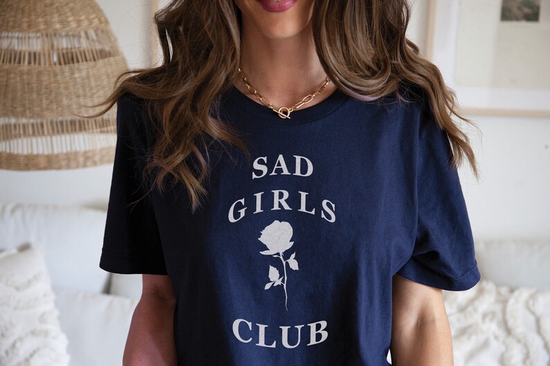 May include: A navy blue t-shirt with the text "SAD GIRLS CLUB" in white lettering and a white rose graphic.