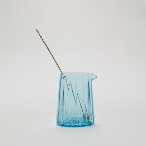 May include: A light blue glass pitcher with a textured surface and a silver stirring rod inside. The pitcher has a spout and a handle.