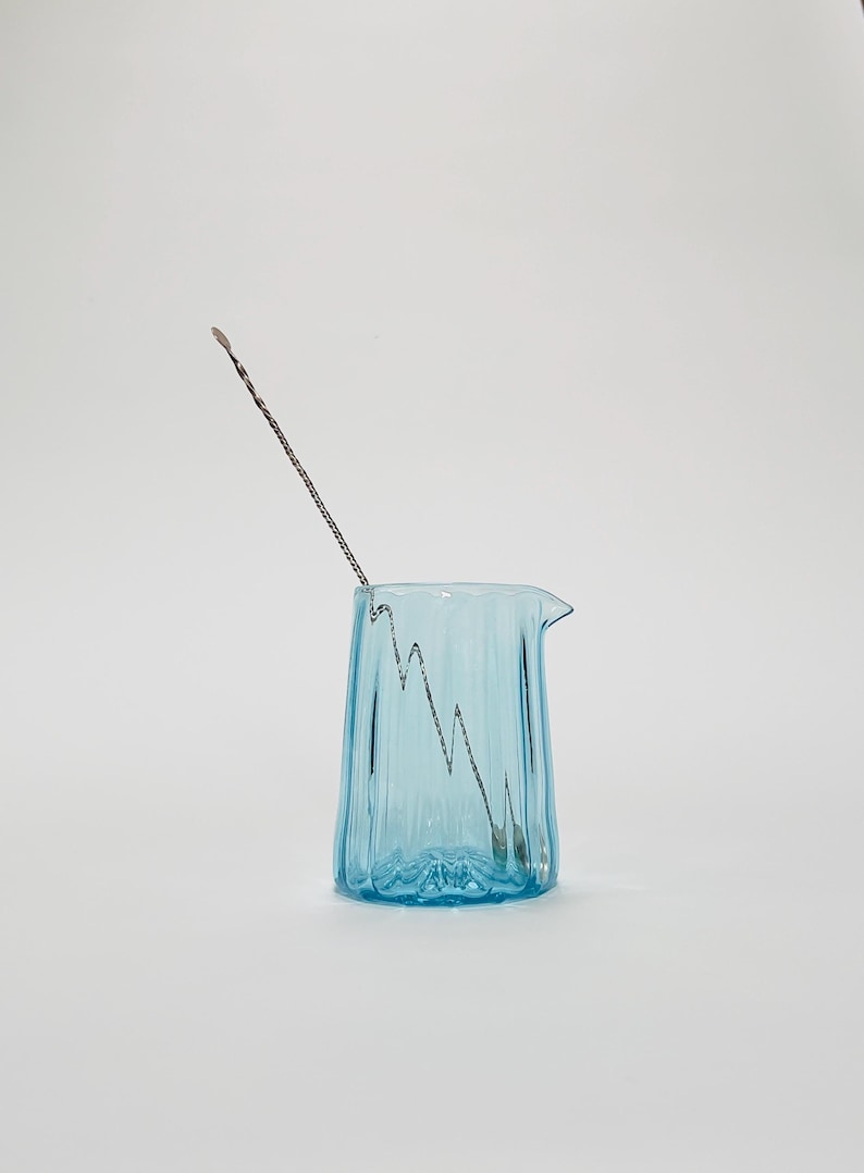 May include: A light blue glass pitcher with a textured surface and a silver stirring rod inside. The pitcher has a spout and a handle.