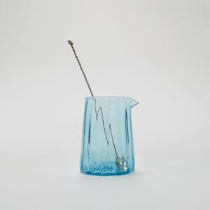 May include: A light blue glass pitcher with a textured surface and a silver stirring spoon inside.