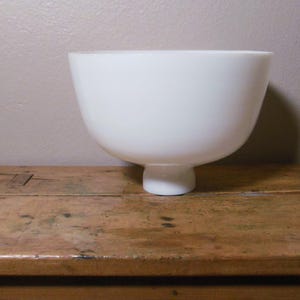 May include: A white, opaque bowl with a rounded shape and a short, pedestal base. The bowl is set against a neutral background and sits on a weathered wooden surface. The bowl's design is simple and elegant.