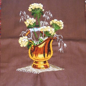 May include: Embroidered artwork featuring a brown pitcher filled with yellow flowers and white accents. The pitcher sits on a white embroidered surface against a brown background. The artwork is labeled "2284" and "Sunset Designs, MCMLXXX".
