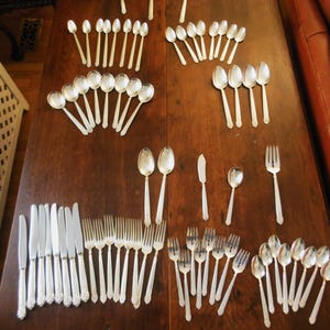 May include: An assortment of silver-colored flatware, including spoons, forks, knives, and serving utensils, arranged on a dark wooden table. The silverware has decorative handles and is displayed in organized sets, ideal for a formal dining setting.
