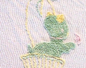 Puede incluir: Un tapiz de pared con textura de punch needle que muestra una rana de color verde claro sentada dentro de una cesta amarilla. La rana está unida a un gran lazo amarillo mediante finos hilos amarillos, y una pequeña flor rosa está a la derecha.