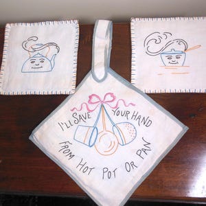 May include: A set of three white embroidered kitchen items. Two square pot holders feature cartoon drawings of a teapot and a pot. A diamond-shaped hot pad with the text "I'll save your hand from hot pot or pan" is in the center.