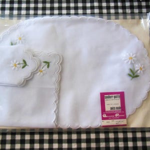 May include: White embroidered placemat and matching napkins set. The placemat is oval-shaped with a scalloped edge and daisy embroidery. The napkins also have daisy embroidery. The set is packaged in a clear plastic bag.