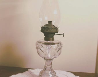 May include: A clear glass oil lamp with a decorative floral pattern on the base and a clear glass chimney. The lamp has a brass-colored burner and a small handle. The lamp is sitting on a white, scalloped-edge doily.