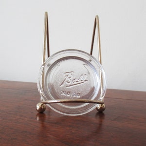 May include: A glass jar lid with the Ball logo in a gold metal stand. The stand has a round top and three legs.