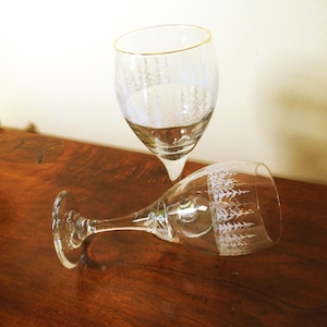 May include: Two clear wine glasses with a gold rim and a white tree pattern. One glass is upright, the other is lying on its side on a dark wooden surface. The glasses are elegant and suitable for serving wine.