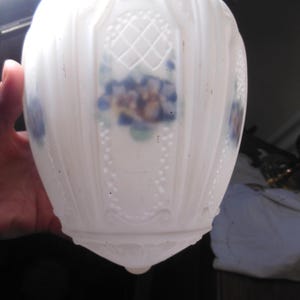 May include: A white, vintage glass lampshade with a floral design. The lampshade has a rounded shape with vertical ribbed detailing and a decorative floral pattern in blue and yellow. The bottom of the lampshade has a pointed, teardrop shape.