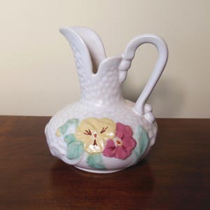 May include: White ceramic pitcher with a textured surface and a handle. The pitcher is decorated with yellow and pink pansies and green leaves. A decorative floral pitcher.