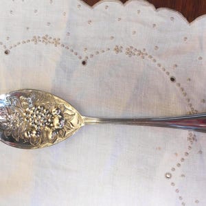 May include: Silver serving spoon with an ornate floral and grape design on the bowl. The spoon is resting on a white, embroidered cloth. The handle has a heart-shaped design at the end.