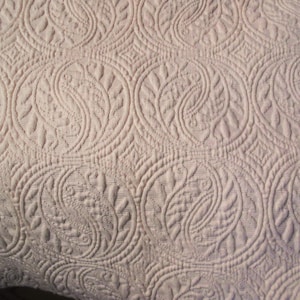 May include: A light lavender textile featuring an intricate, repeating quilted pattern of circular motifs. Each circle contains stylized, curved leaf-like shapes and textured stitching, creating a raised, embossed appearance across the fabric surface.