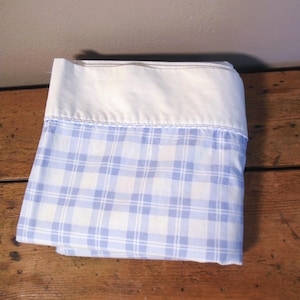 May include: Folded white and blue plaid bed sheet. The top portion is solid white, while the bottom features a blue and white plaid pattern. The sheet is neatly folded, showcasing the contrasting colors and textures. The fabric appears to be cotton or a similar material.