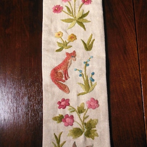 May include: A long, rectangular strip of off-white linen fabric featuring detailed embroidery of a fox, a bird, and various colorful flowers including pink, yellow, and purple blossoms, along with green leaves and stems arranged in a vertical pattern.