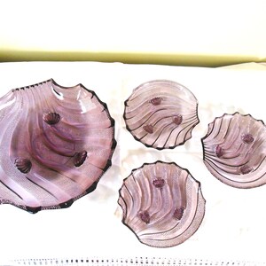 May include: A set of four purple glass dishes featuring a shell-inspired design with scalloped edges and textured, ribbed surfaces. The collection includes one large serving bowl and three smaller matching plates, all with a translucent finish.