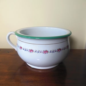 May include: White ceramic chamber pot with a green rim and handle. A floral pattern of pink and blue flowers decorates the exterior. The pot sits on a dark wooden surface.