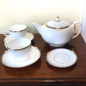 May include: A white porcelain tea set with gold and navy blue trim. The set includes a teapot, two teacups, and saucers. The teapot has a gold finial on the lid and a curved handle. The teacups and saucers have a matching design.