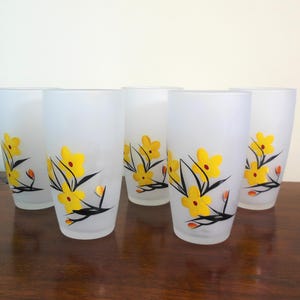 May include: Set of five frosted glass tumblers. Each tumbler features a yellow floral design with black stems and orange accents. The glasses are cylindrical and sit on a dark wooden surface.