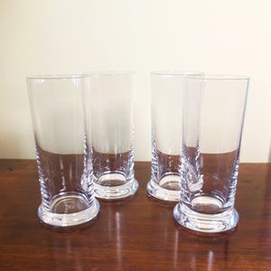 May include: Four clear, cylindrical drinking glasses with a rounded base are arranged on a dark wooden surface. The glasses are empty and reflect the light, showcasing their transparent nature. The background is a plain, light-colored wall.