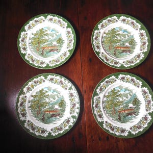 May include: Four round ceramic plates featuring a central landscape illustration of trees, a small building, and a body of water. The rims are decorated with a green textured border and a repeating pattern of small yellow and red floral clusters.