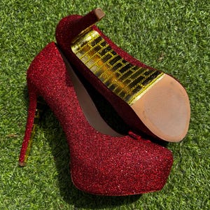 Könnte beinhalten: Ein Paar rote Glitzer-Plateau-High Heels. Die Schuhe haben einen Stiletto-Absatz und eine Plateausohle. Die Innensohle ist goldfarben. Die Schuhe liegen auf grünem Gras.