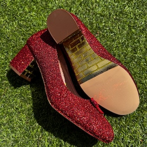 May include: A pair of red glitter heels with a low block heel. The shoes have a rounded toe and a small bow detail. The soles are a light brown colour with a gold brick pattern on the bottom.