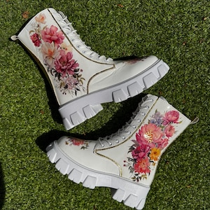 May include: White combat boots with a floral pattern. The boots have a chunky sole, lace-up fastening, and are decorated with pink, red, and yellow flowers. Gold trim accents the edges, adding a touch of elegance. The boots are a stylish footwear choice.