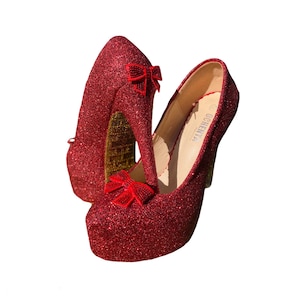 Könnte beinhalten: Ein Paar rote Glitzer-High Heels mit roten Schleifen an der Vorderseite. Die Absätze sind etwa 12,7 cm hoch und haben eine Plattform.
