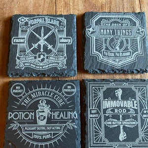 May include: Four black slate coasters with white designs. The coasters feature illustrations of a sword, a deck of cards, a potion bottle, and a rod. The text on the coasters reads "The Vorpal Blade", "The Deck of Many Things", "The Miracle Cure Potion of Healing", and "Immovable Rod".