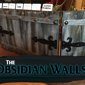 Puede incluir: Un tablero de juego de madera con un acabado gris y negro desgastado. El tablero está dividido en secciones con bisagras de metal negro y detalles decorativos. El tablero está etiquetado como "The Obsidian Walls".