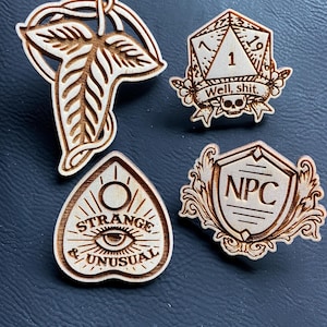 May include: Four wooden pins with different designs. The first pin is a leaf with a stem. The second pin is a 20-sided die with the number 1 showing and the text "Well, shit." The third pin is a heart-shaped Ouija board with the text "Strange & Unusual." The fourth pin is a shield with the text "NPC."