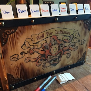 May include: A wooden box with a black metal trim and brass accents. The box has a label that says "Roll For Initiative" and features a multi-eyed monster with a toothy grin. There are white cards with names written on them, such as "Vax", "Percy", "Scanlan", "Vecna", "Grog", "Death Knight", "Vex", "Kehlah", and "Pike".
