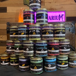 May include: A collection of 15 candles in glass jars with gold lids. Each candle has a different label with a unique design and name. The candles are arranged in a pyramid shape on a dark surface. The labels include names like "Mirkwood", "Forbidden Forest", "Yggdrasil", and "The Shire".