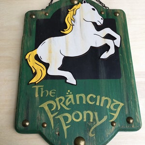 Puede incluir: Un letrero de madera con fondo verde y letras doradas que dice "The Prancing Pony". Un caballo blanco con una crin y cola amarillas está representado en el letrero.