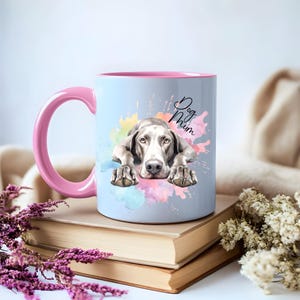 Puede incluir: Una taza de cerámica azul claro con un borde y un asa de color rosa. La taza presenta una ilustración de acuarela de un perro Weimaraner con el texto "Dog Mum" en negro.