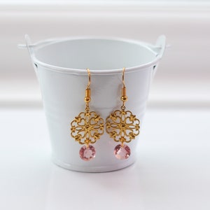 May include: Gold filigree earrings with pink crystal drops. The earrings are hanging from a white bucket.