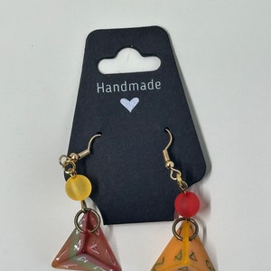 Può includere: Orecchini fatti a mano con un ciondolo a forma di dado triangolare di colore rosso fumè e arancione con dettagli dorati. Gli orecchini sono esposti su una carta nera con la scritta "Handmade" e un cuore.